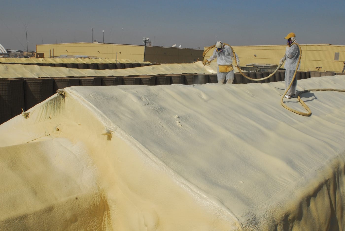 Applicators in protective suits spraying polyurethane foam across a large commercial roof surface